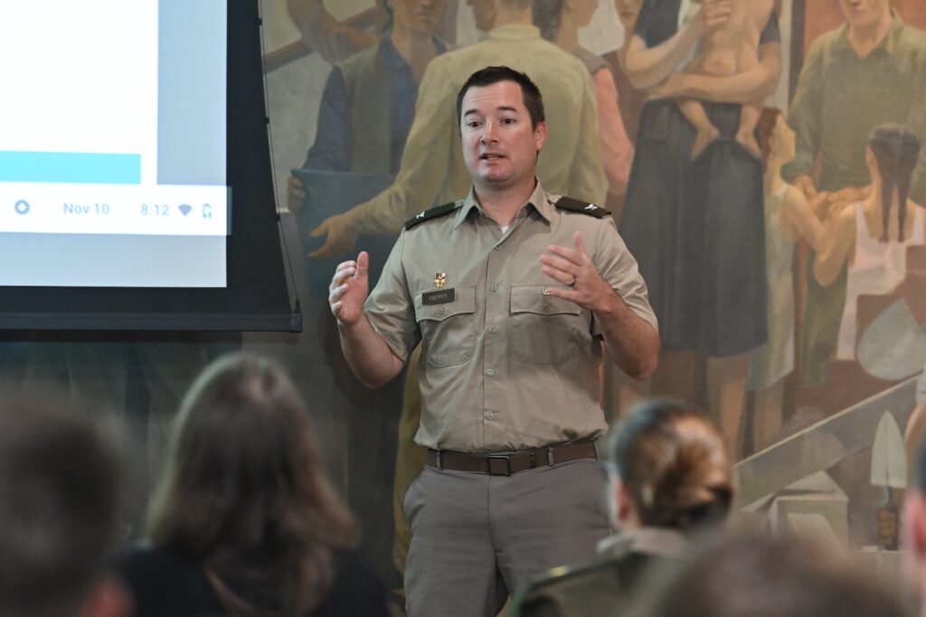 Knepper stands in front of a screen and mural while addressing cadets.