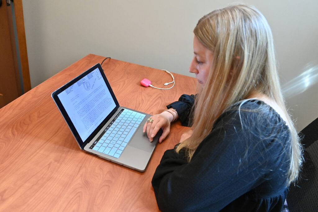 A college student sits at a desk and works on her laptop while conducting research.